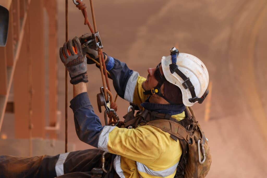 A technician in high-visibility gear and a hard hat carefully adjusting rope access rigging and safety lines at a Melbourne industrial site.