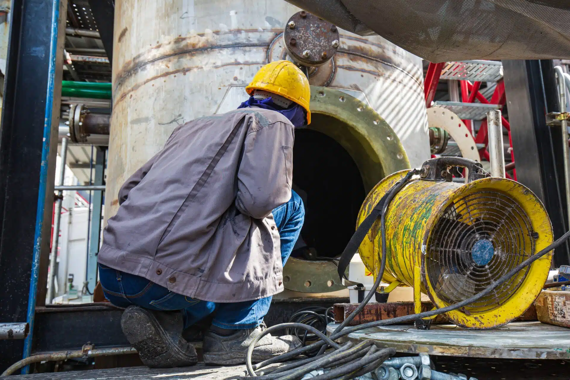 An industrial worker monitoring a large storage vessel with a yellow forced-air ventilation fan in the foreground, demonstrating confined space hazard controls.