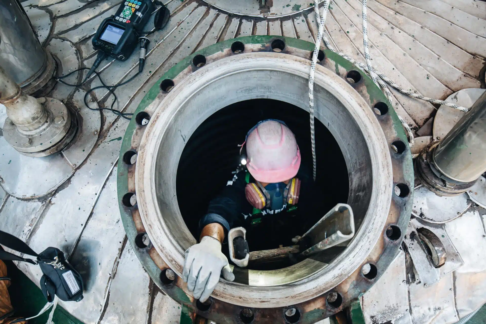 High-angle view of a worker using a ladder and breathing apparatus to exit a circular manhole, with atmospheric monitoring equipment visible on deck.
