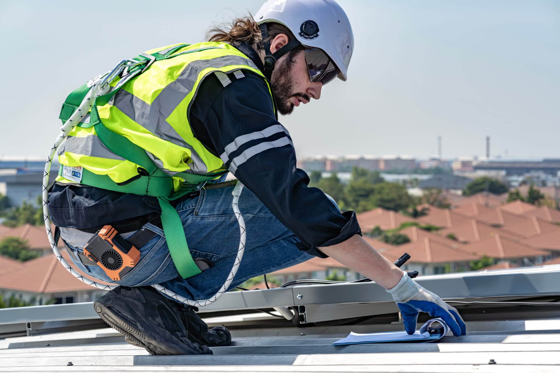 A professional in a safety harness and high-visibility vest reviewing site plans while working on a metal roof, demonstrating working at heights compliance.