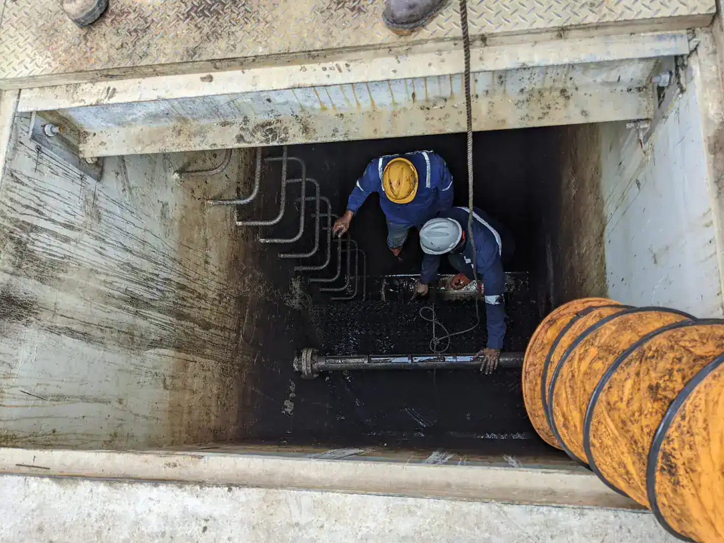 A high-angle view looking down into a deep, muddy industrial pit where two workers in coveralls and hard hats are conducting a confined space entry.