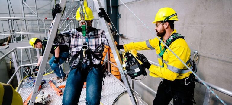 Three workers in yellow hard hats and safety gear are working at height, possibly using ropes access techniques.