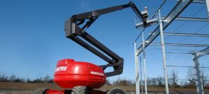 A red Manitou articulated boom lift with a worker in the platform is positioned near steel beams against a blue sky.